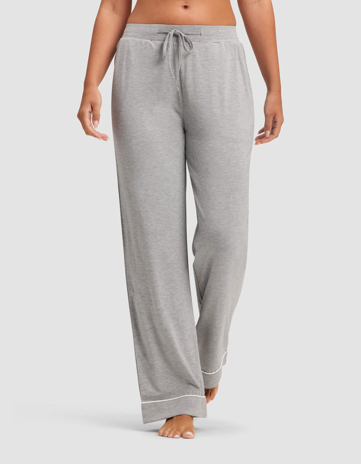 A woman wears Cozy Earth Women's Bamboo Stretch-Knit Pajama Pants in light gray, featuring a drawstring waist and white trim at the cuffs, standing barefoot against a plain background.|Color:Grey