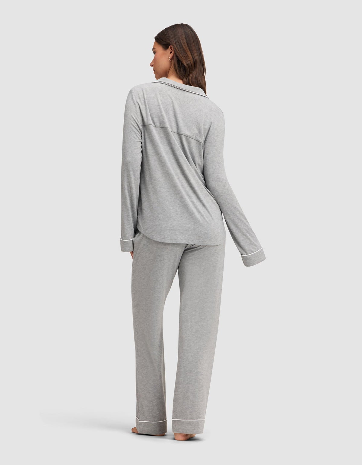 A woman stands barefoot, facing away, wearing the Cozy Earth Women's Bamboo Stretch-Knit Long Sleeve Pajama Set in light gray with white trim. She is on a plain light background. |Color:Grey