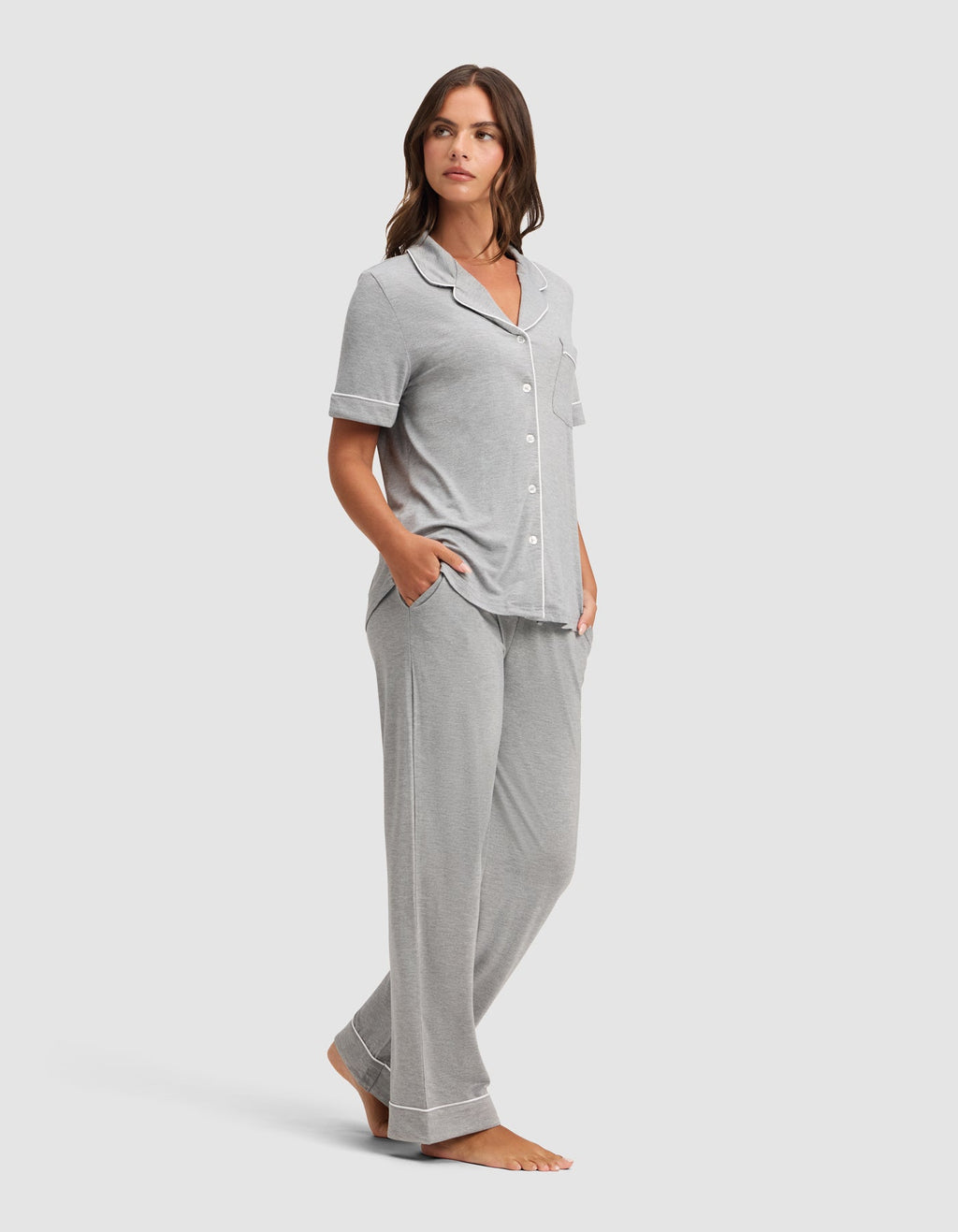 A woman stands barefoot in a Cozy Earth Women's Bamboo Stretch-Knit Short Sleeve & Pant Pajama Set, featuring light gray fabric with white piping and loose pants, hands in pockets, against a plain light background.  |Color:Grey