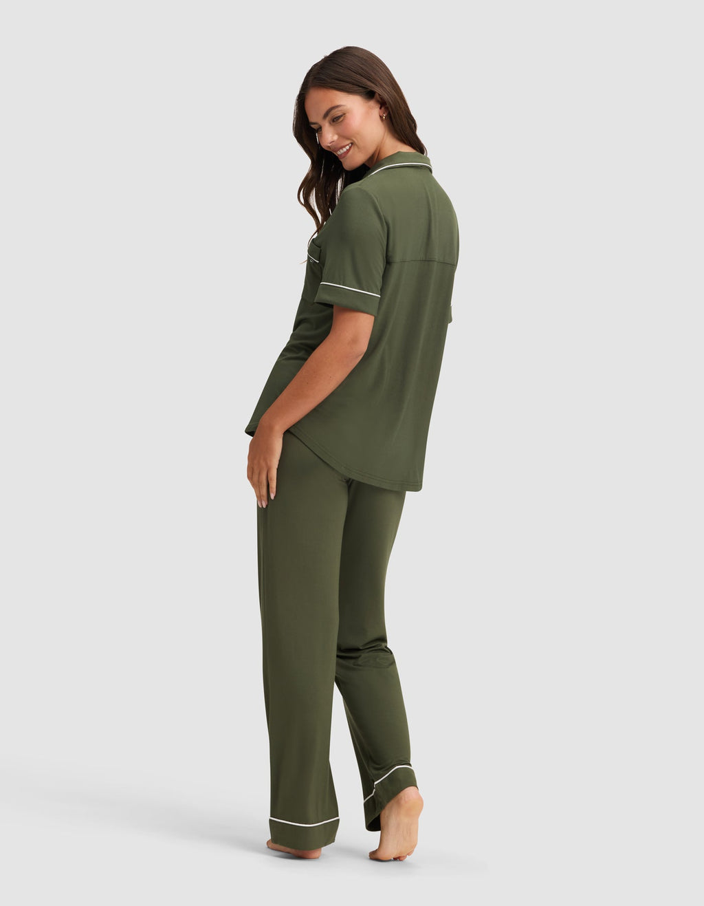 A woman stands barefoot, smiling over her shoulder, wearing the Cozy Earth Women's Bamboo Stretch-Knit Short Sleeve & Pant Pajama Set in olive green with white piping. The background is plain light gray.|Color:Olive