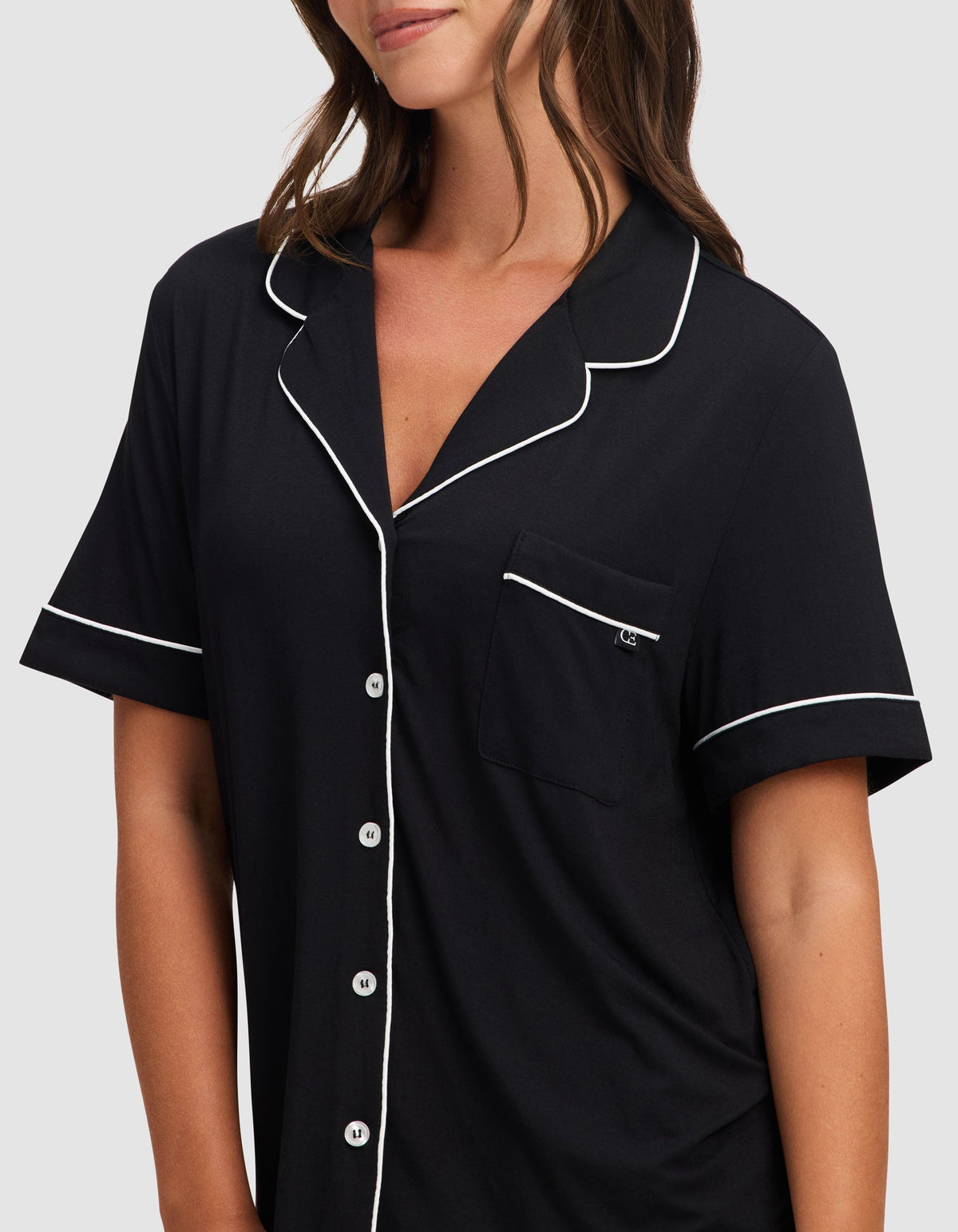 A woman wears the Cozy Earth Women's Bamboo Stretch-Knit Short Sleeve Pajama Set in black, featuring white piping, front buttons, and a chest pocket, standing against a plain light background.  |Color:Black