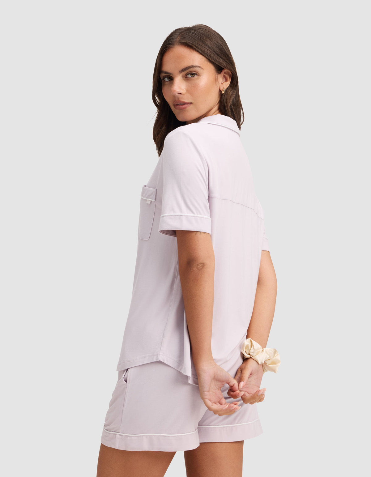 A woman models the Cozy Earth Women's Bamboo Stretch-Knit Short Sleeve Pajama Set in light lavender, standing with her back slightly turned, hands clasped behind her, and a beige scrunchie on her wrist. |Color:Lilac