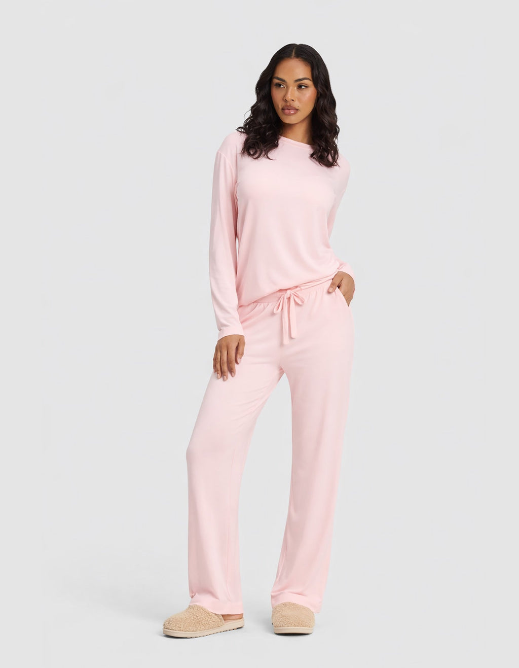A woman wears the Cozy Earth Women's Bamboo Stretch-Knit Long Sleeve Lounge Tee & Pant Set in Blossom, paired with beige slippers, standing against a plain light gray background.|Color:Blossom
