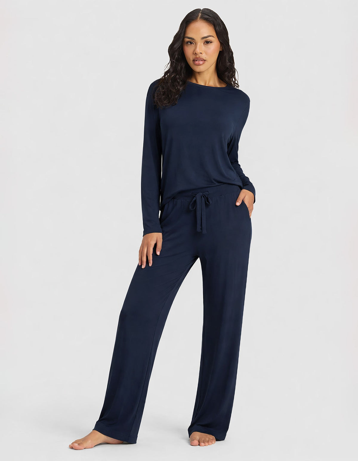 A woman poses barefoot in Cozy Earth's Women's Bamboo Stretch-Knit Long Sleeve Lounge Tee & Pant Set in navy, standing confidently against a plain light background.|Color:Navy