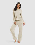 A woman with long, wavy dark hair models the Cozy Earth Women's Loews Hotel Lounge Set—a loose, light beige pajama set with long sleeves and wide-leg pants—as she stands barefoot against a plain white background.|Color:Warm Grey