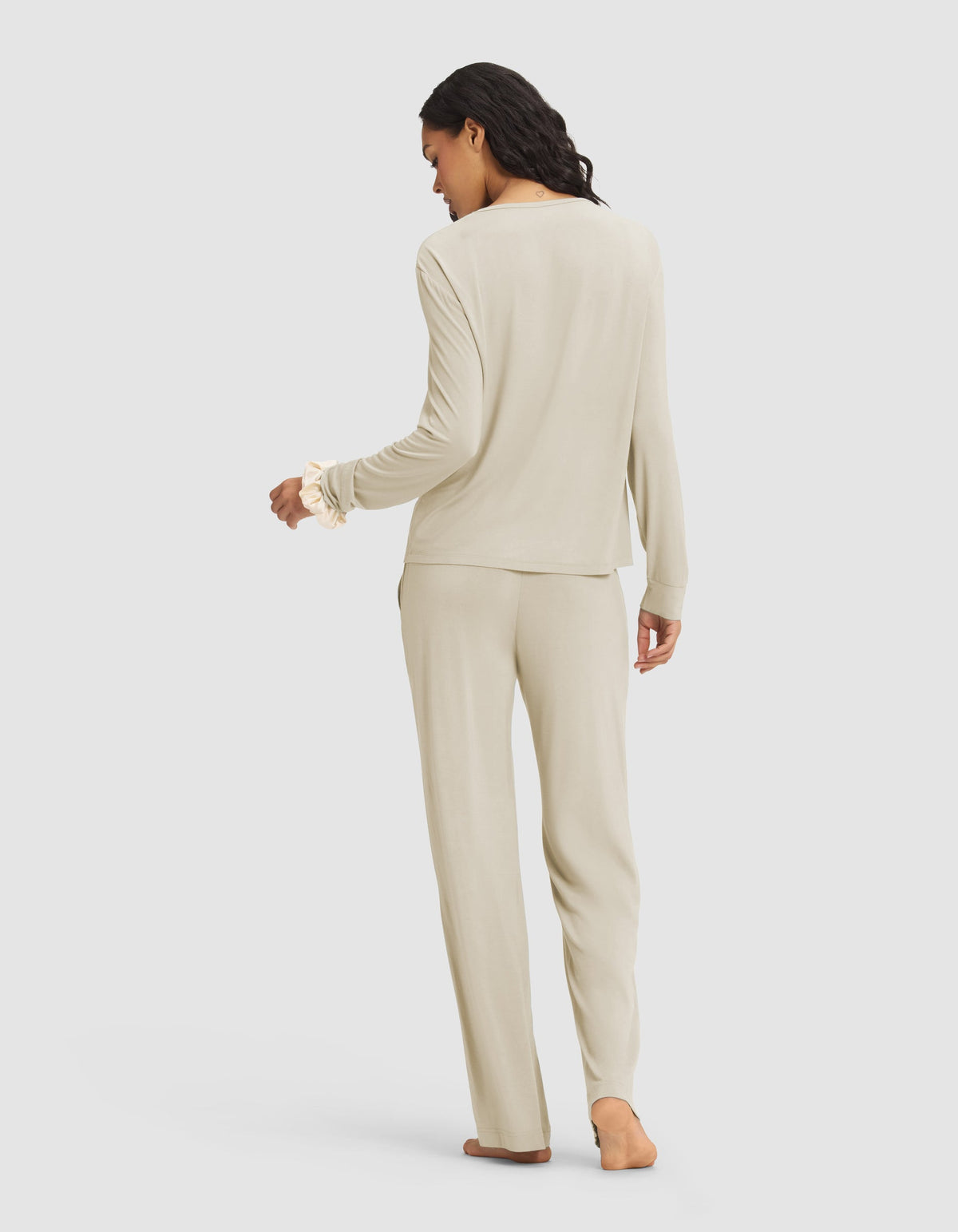 A woman with dark, wavy hair stands barefoot and facing away in the Cozy Earth Women's Loews Hotel Lounge Set—a light beige long-sleeve pajama top with matching pants—against a plain light gray background.|Color:Warm Grey