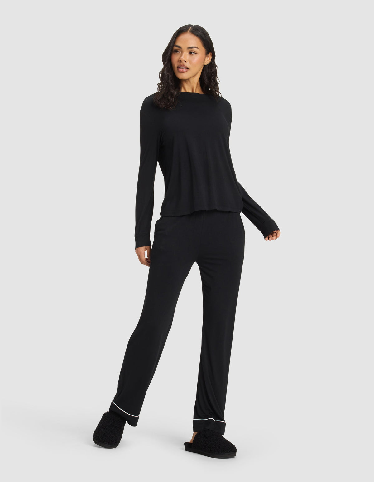 A woman stands against a plain background wearing the Cozy Earth Women's Bamboo Stretch-Knit Long Sleeve Lounge Tee in black and matching slippers, smiling slightly with one hand on her hip and looking to the side.|Color:Black