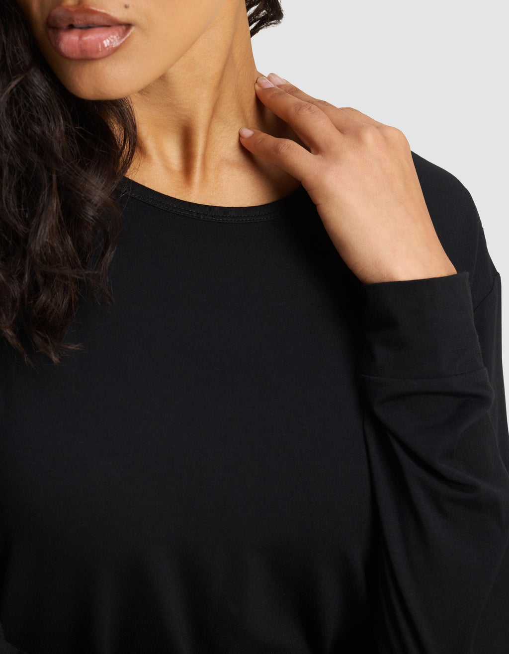 A woman with wavy dark hair wears the Women's Bamboo Stretch-Knit Long Sleeve Lounge Tee by Cozy Earth. She gently touches her neck with her right hand, and only the lower half of her face and upper torso are visible.|Color:Black