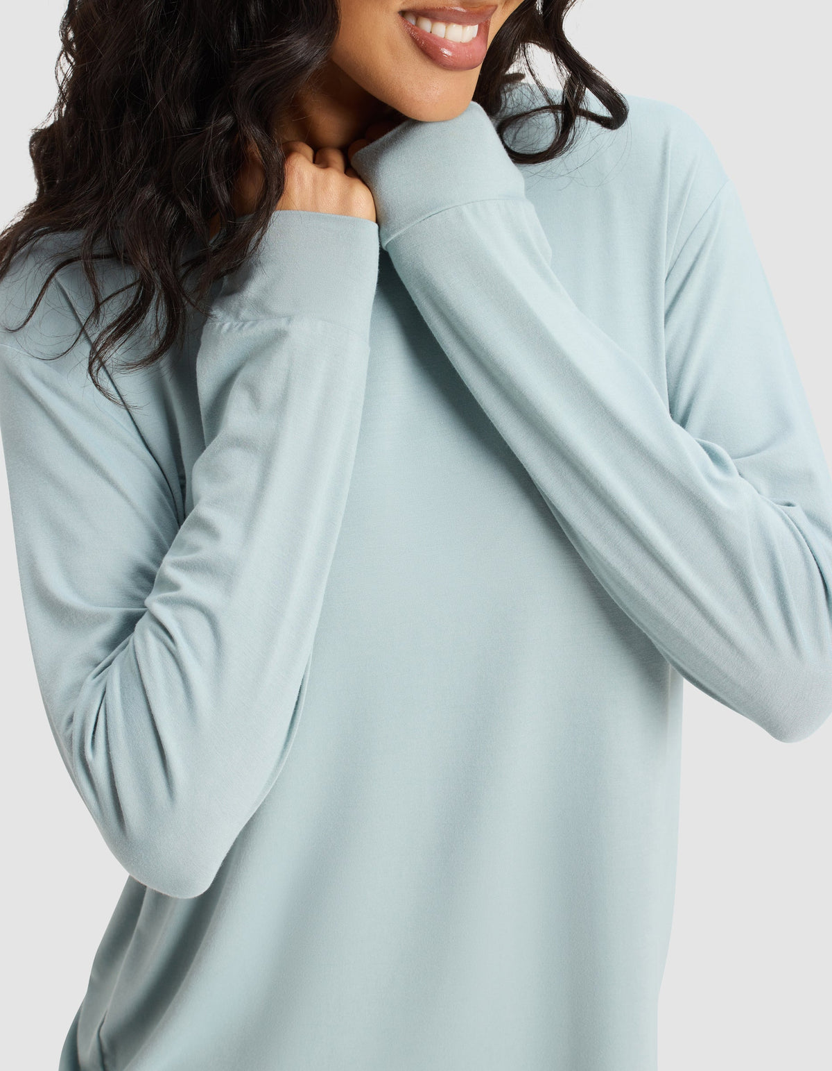 A woman with wavy dark hair smiles while holding the collar of her light blue Women's Bamboo Stretch-Knit Long Sleeve Lounge Tee by Cozy Earth, her face partially visible against a plain background.|Color:Brook