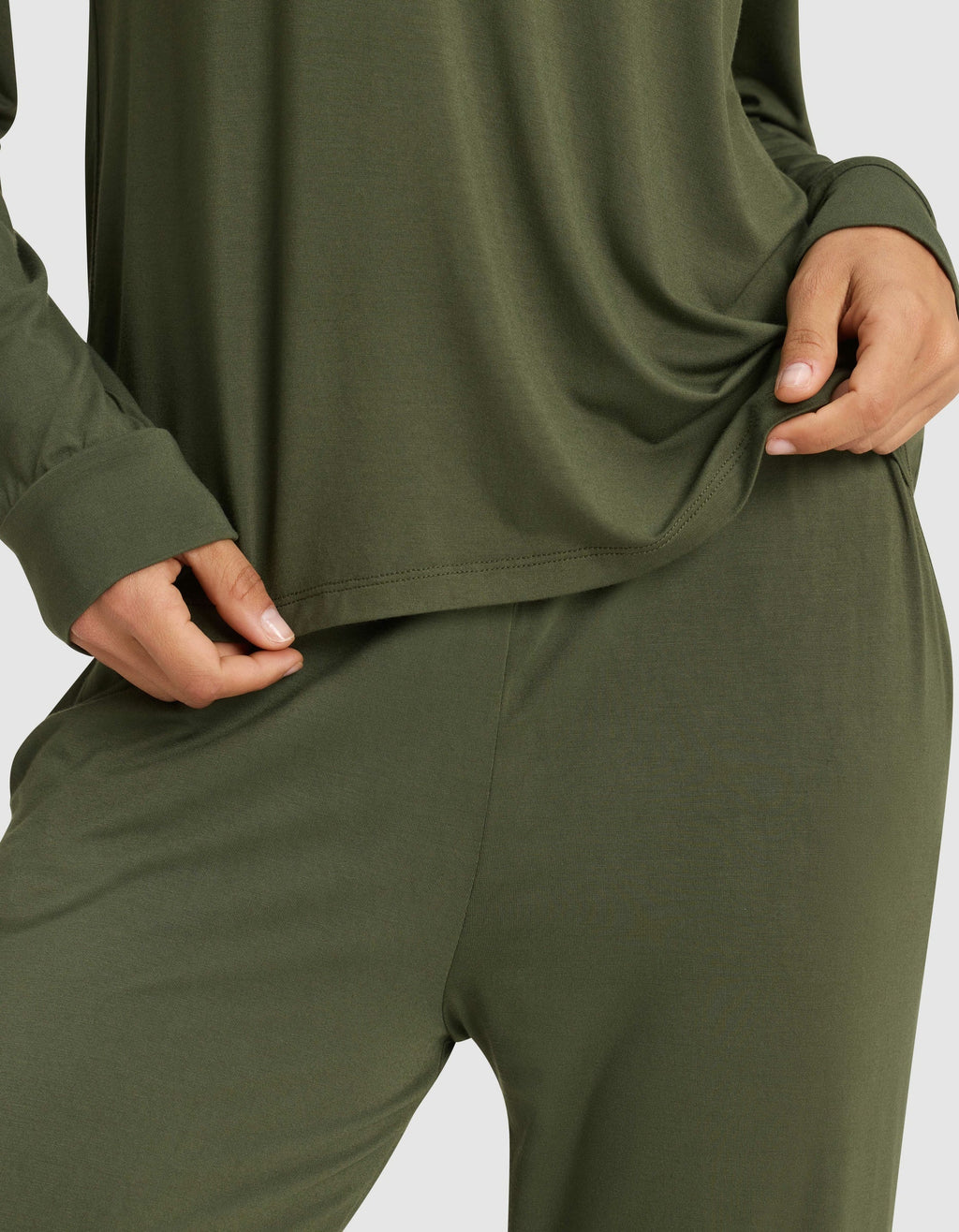 A woman wears the Cozy Earth Women's Bamboo Stretch-Knit Long Sleeve Lounge Tee in olive green, holding the shirt's hem; only her torso and upper legs are visible.|Color:Olive