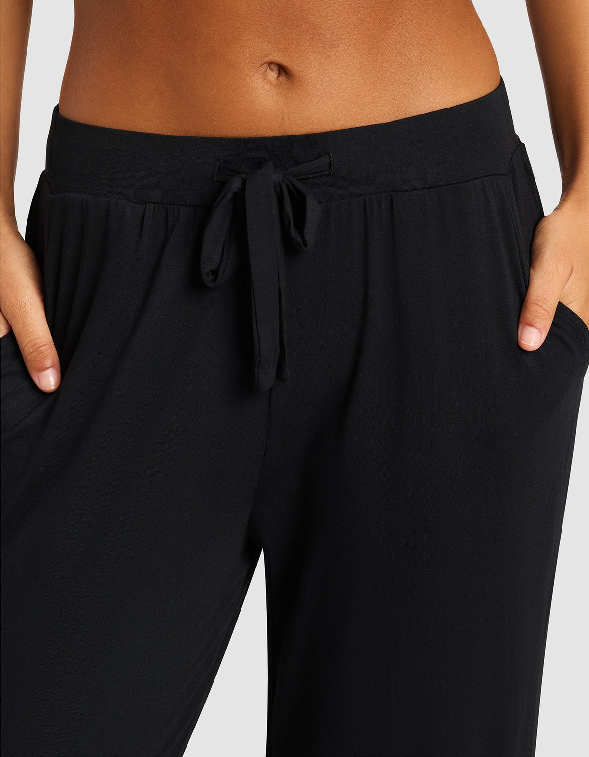 A close-up shows a person wearing Cozy Earth's Women's Bamboo Stretch-Knit Pant, hands in the pockets, highlighting the waistband and midriff against a plain background.|Color:Black