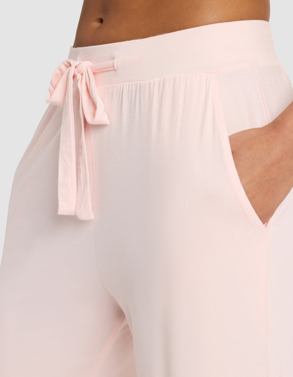 A woman wears Cozy Earth's Women's Bamboo Stretch-Knit Pant in light pink, featuring a drawstring bow at the waist and her hand in the side pocket.|Color:Blossom