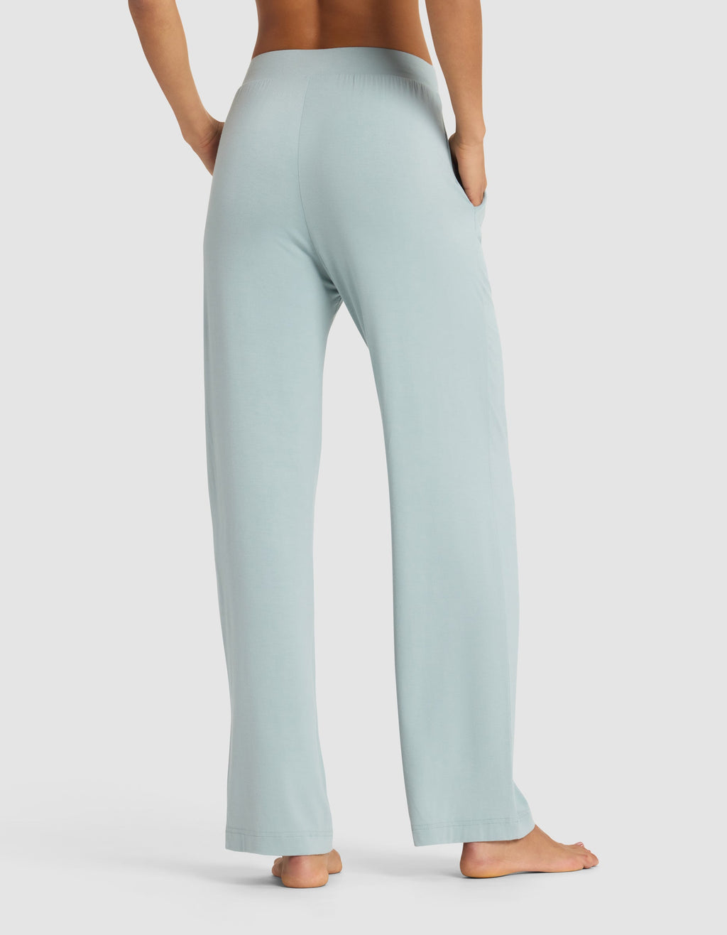 A woman stands barefoot, viewed from behind, wearing Cozy Earth’s Women's Bamboo Stretch-Knit Pant in light blue with hands in the side pockets. The background is plain white.|Color:Brook