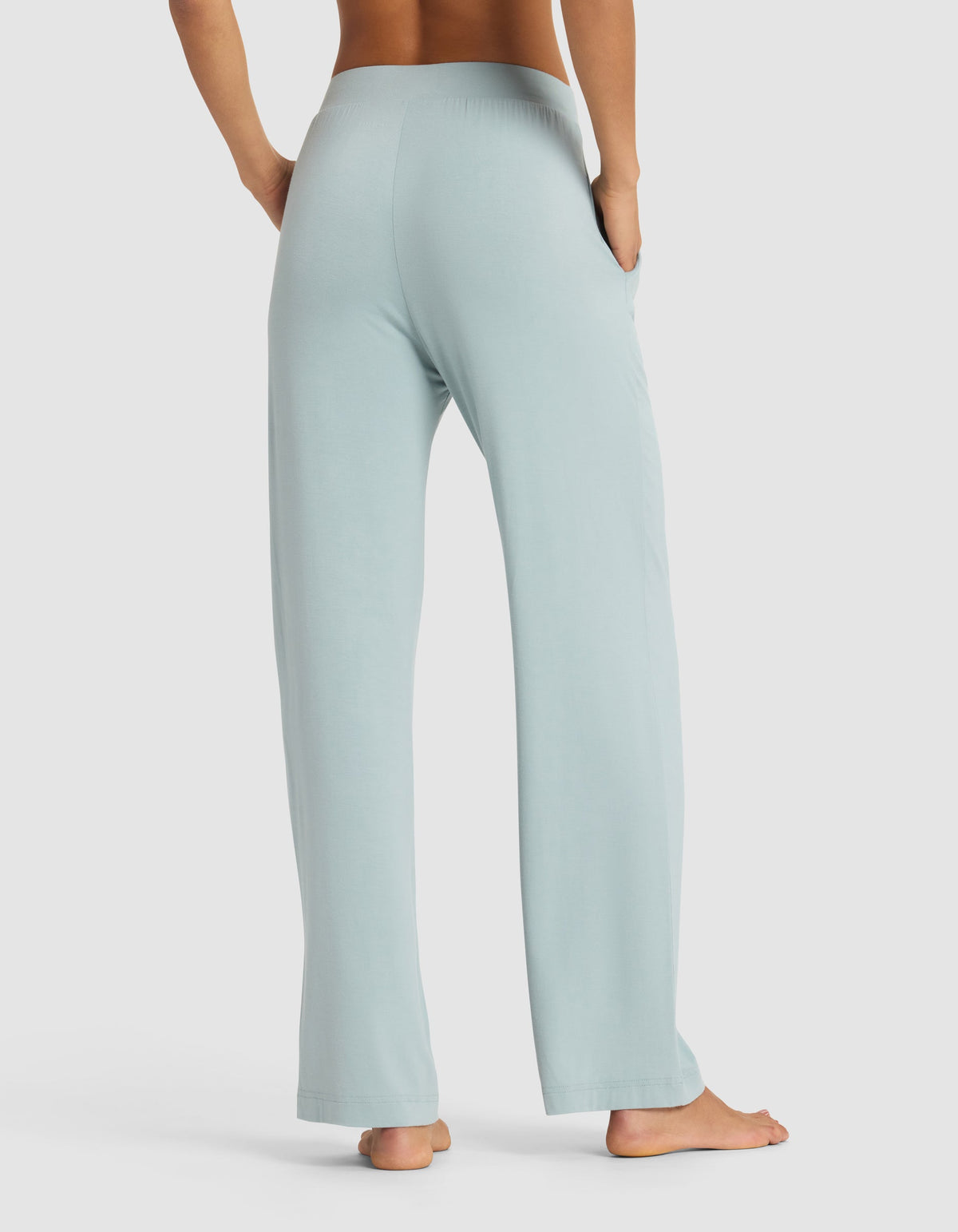 A woman stands barefoot, viewed from behind, wearing Cozy Earth’s Women's Bamboo Stretch-Knit Pant in light blue with hands in the side pockets. The background is plain white.|Color:Brook