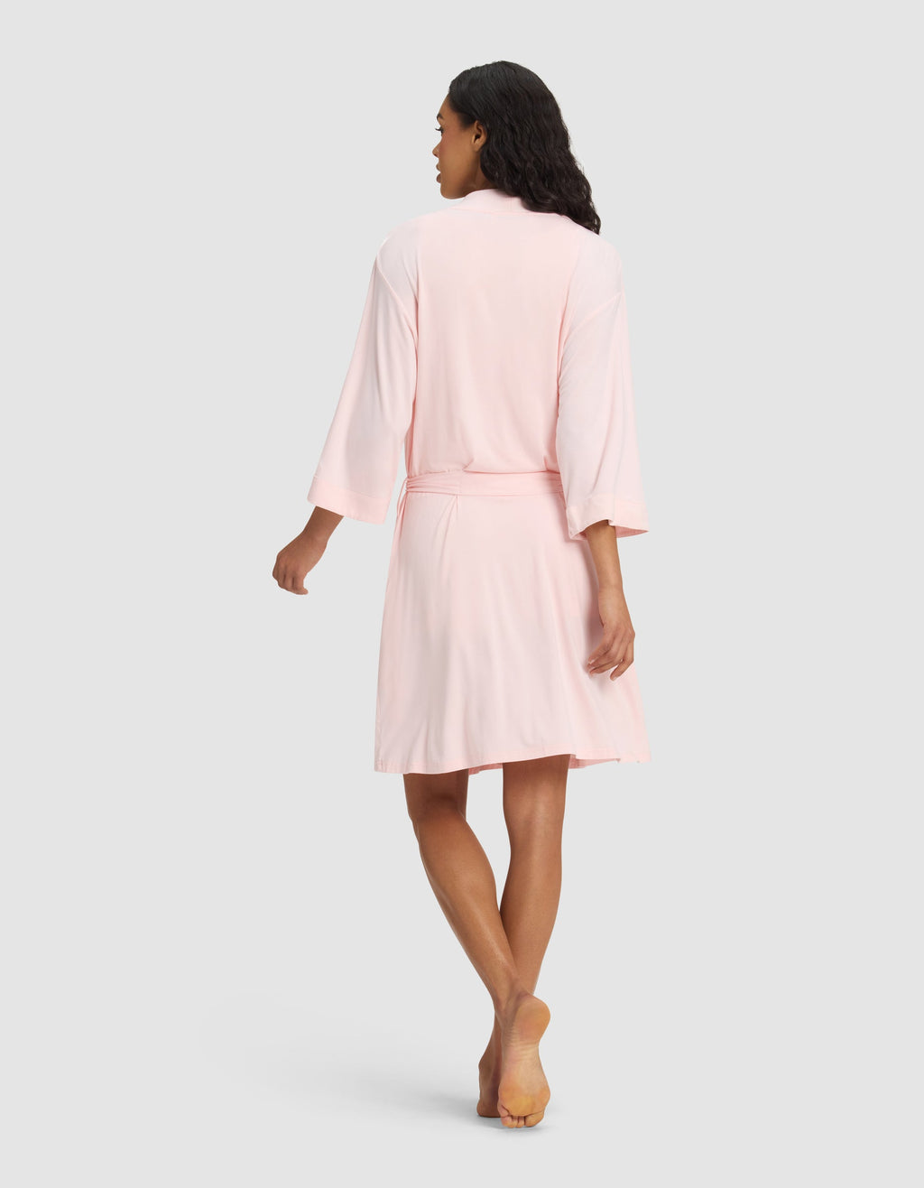 A woman models the Cozy Earth Women's Bamboo Stretch-Knit Kimono Robe in light pink, barefoot on a plain light gray background, shown from the back with her head turned slightly to the side.|Color:Blossom