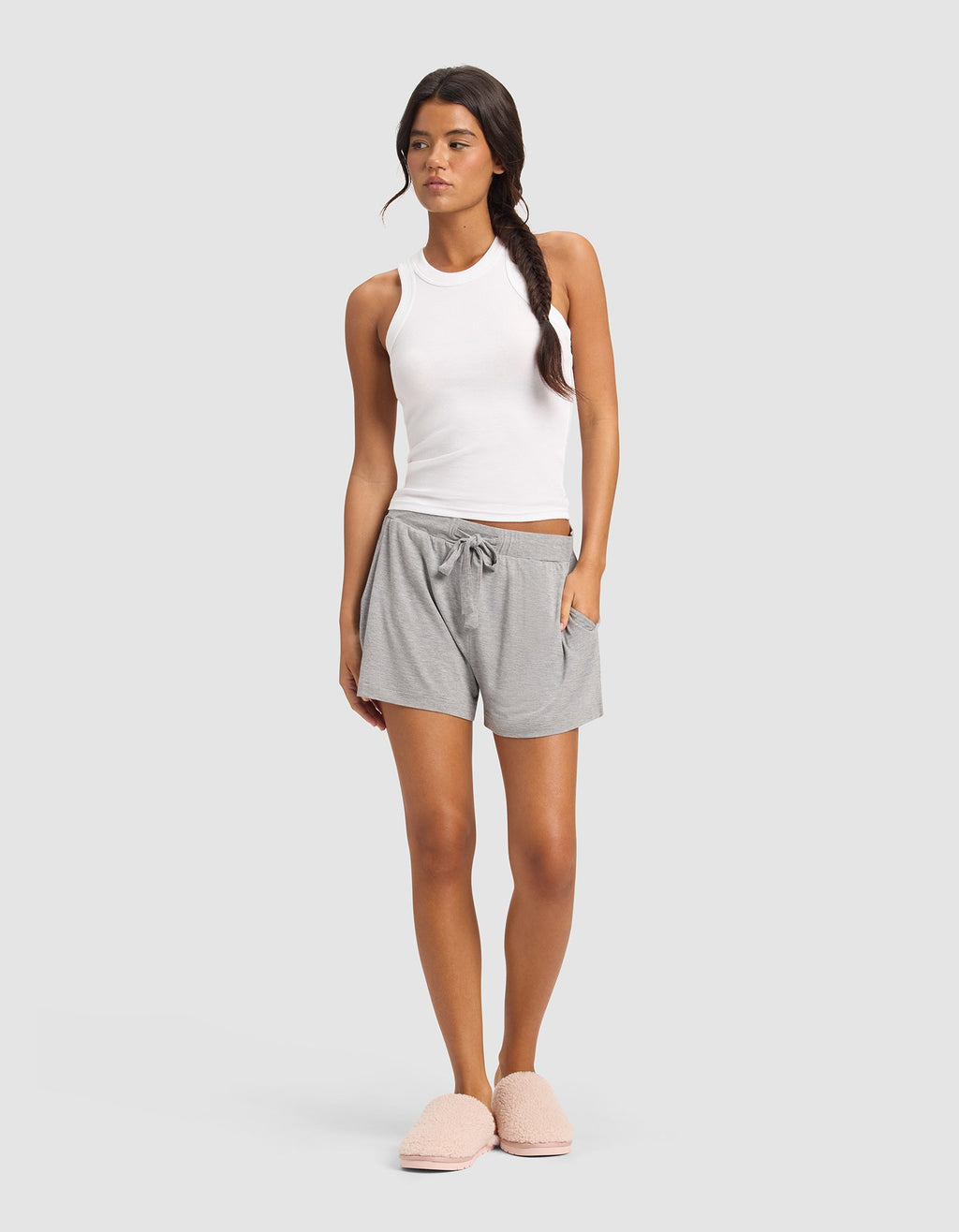 A woman stands against a plain background wearing Cozy Earth's Women's Bamboo Stretch-Knit Shorts in light gray, paired with a white sleeveless tank top and pink fuzzy slippers. Her hair is styled in a loose side braid.|Color:Heather Grey