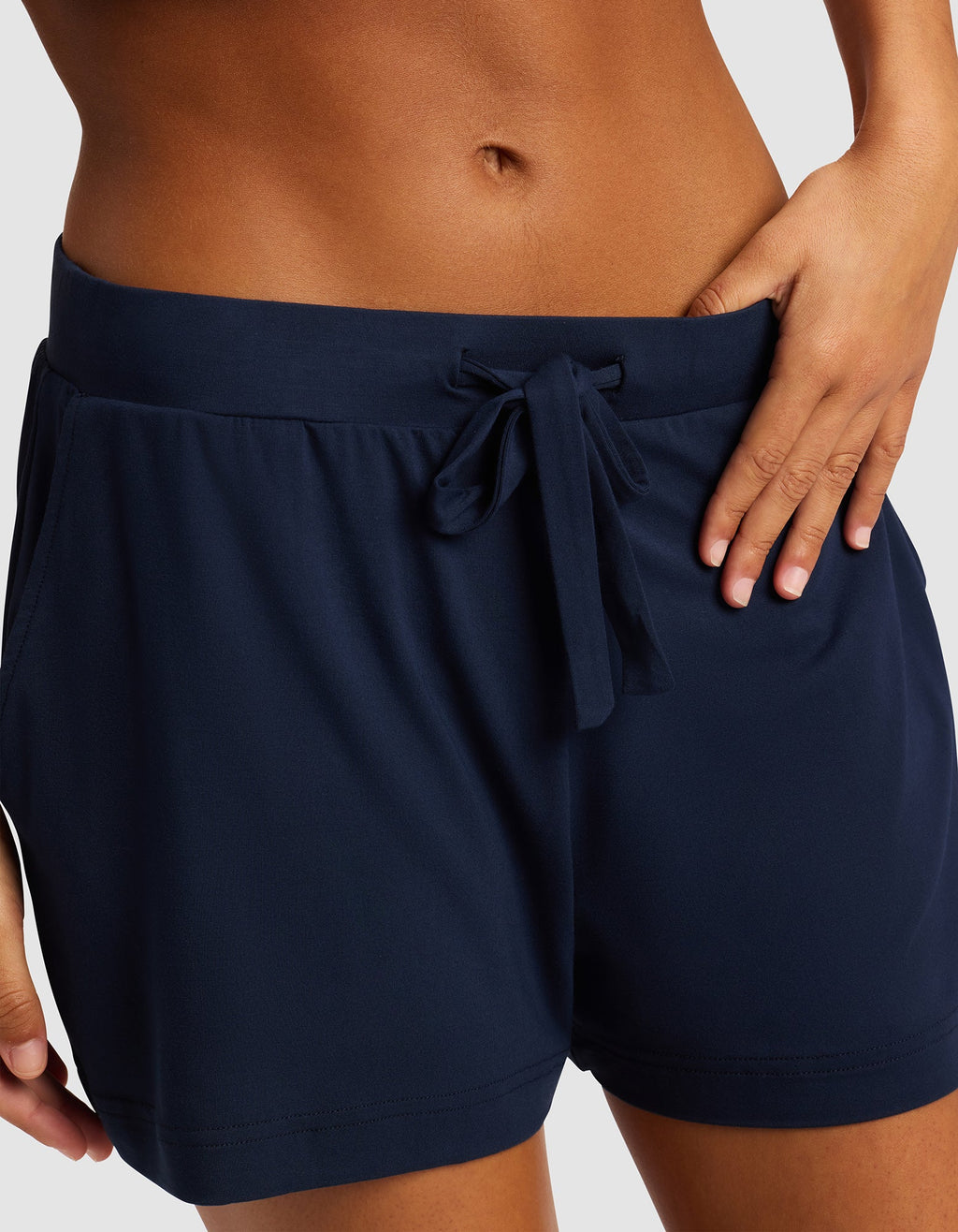 A woman wears Cozy Earth Women's Bamboo Stretch-Knit Shorts in navy blue, featuring a drawstring waistband and side pockets, standing with one hand on her hip against a plain background.|Color:Navy