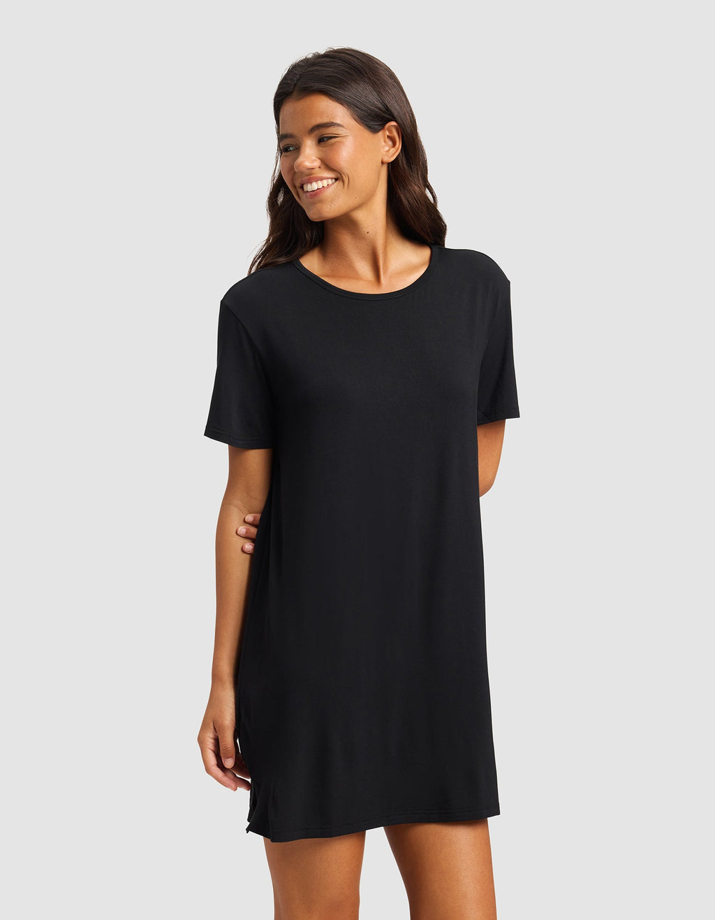 A woman with long dark hair smiles while wearing the Cozy Earth Women's Bamboo Stretch-Knit Sleep Dress, a simple black short-sleeved dress that falls above the knees, standing against a plain light gray background.|Color:Black