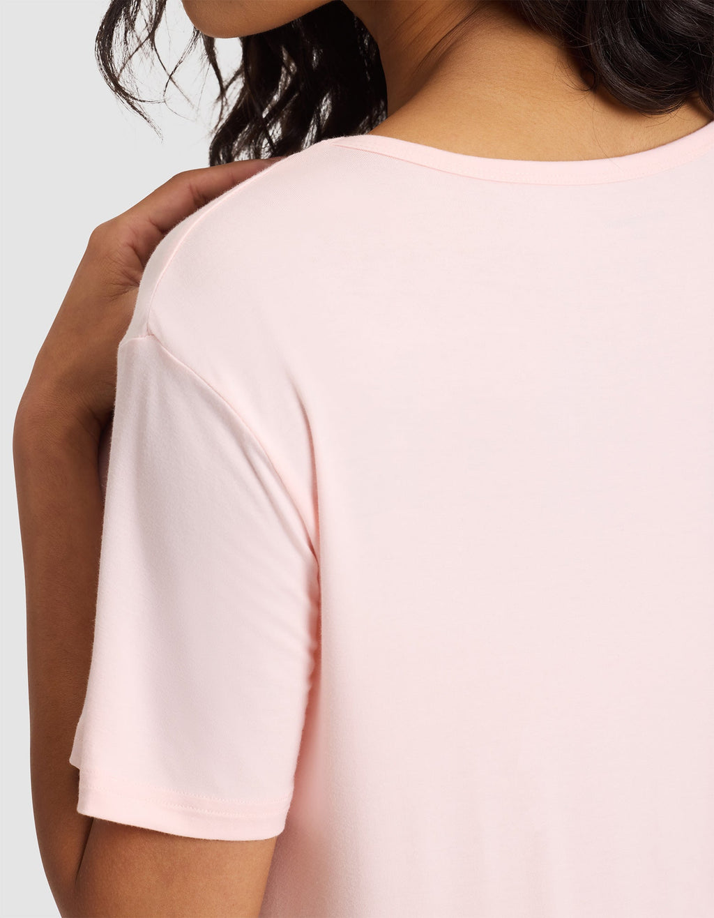 A woman with wavy dark hair models the Cozy Earth Women's Bamboo Stretch-Knit Sleep Dress in light pink, highlighting her shoulder and upper arm against a plain gray background. |Color:Blossom