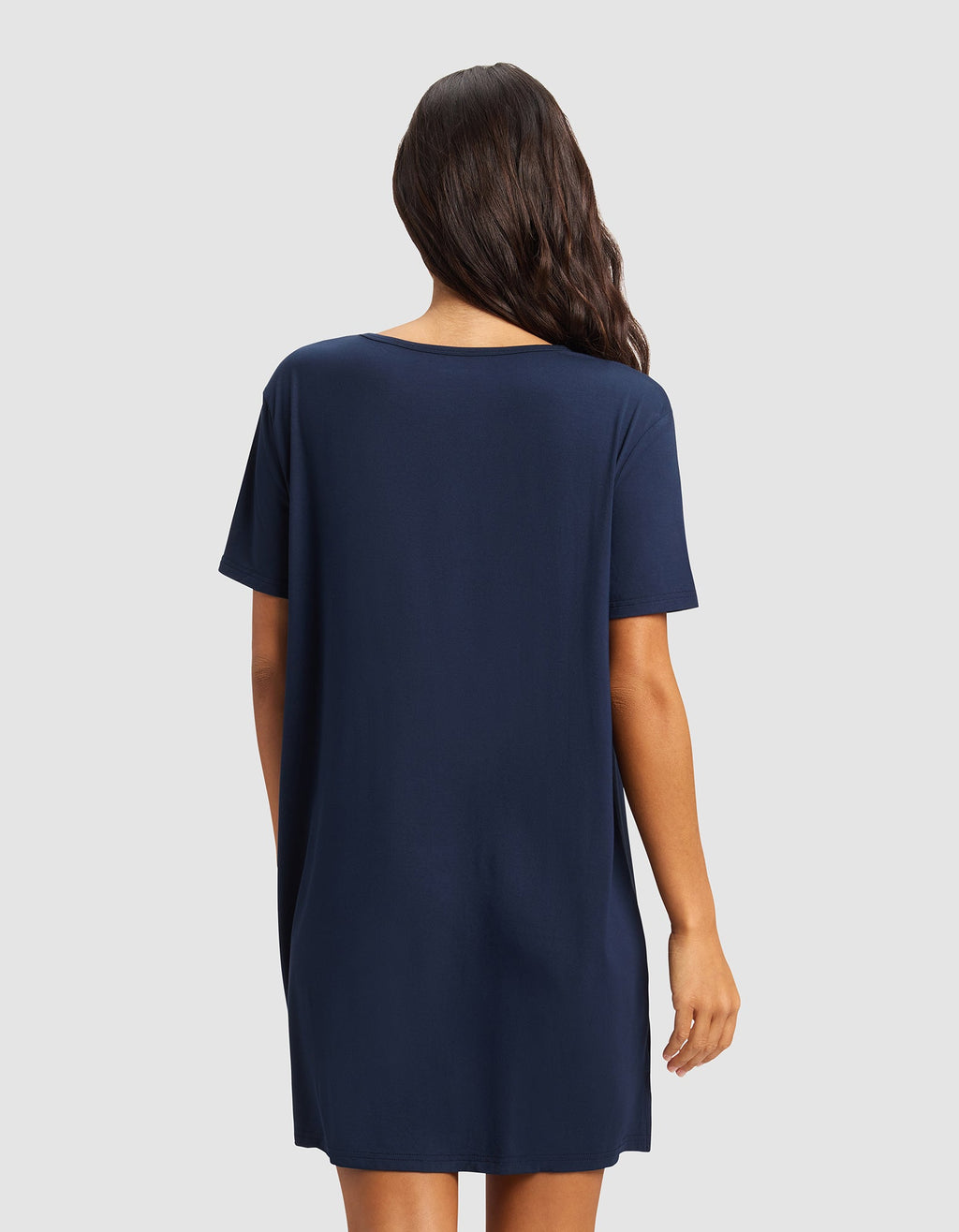 A woman with long wavy hair stands facing away, wearing the Cozy Earth Women's Bamboo Stretch-Knit Sleep Dress in navy blue against a plain light gray background.|Color:Navy