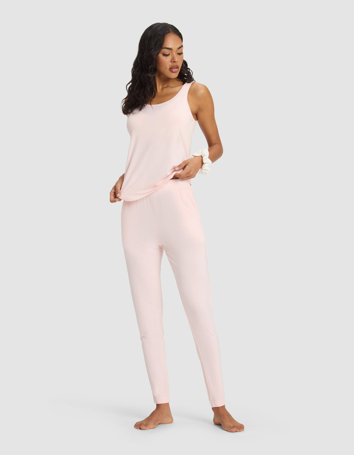 A woman stands barefoot against a plain background, wearing the Cozy Earth Women's Bamboo Stretch-Knit Lounge Tank in light pink with fitted pants. She has long, wavy hair and holds the hem of her top with one hand.|Color:Blossom