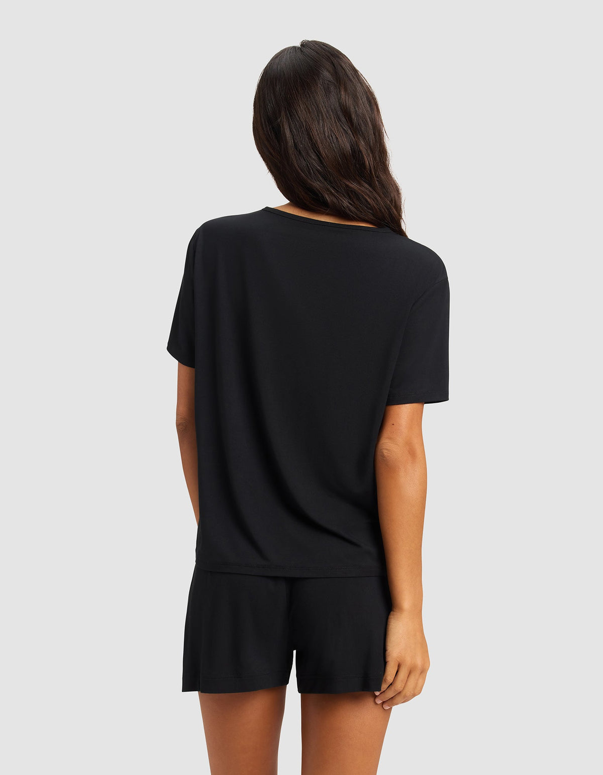 A woman with long brown hair stands with her back to the camera, wearing the Cozy Earth Women's Bamboo Stretch-Knit Lounge Tee in black, paired with matching shorts, against a plain light gray background.|Color:Black
