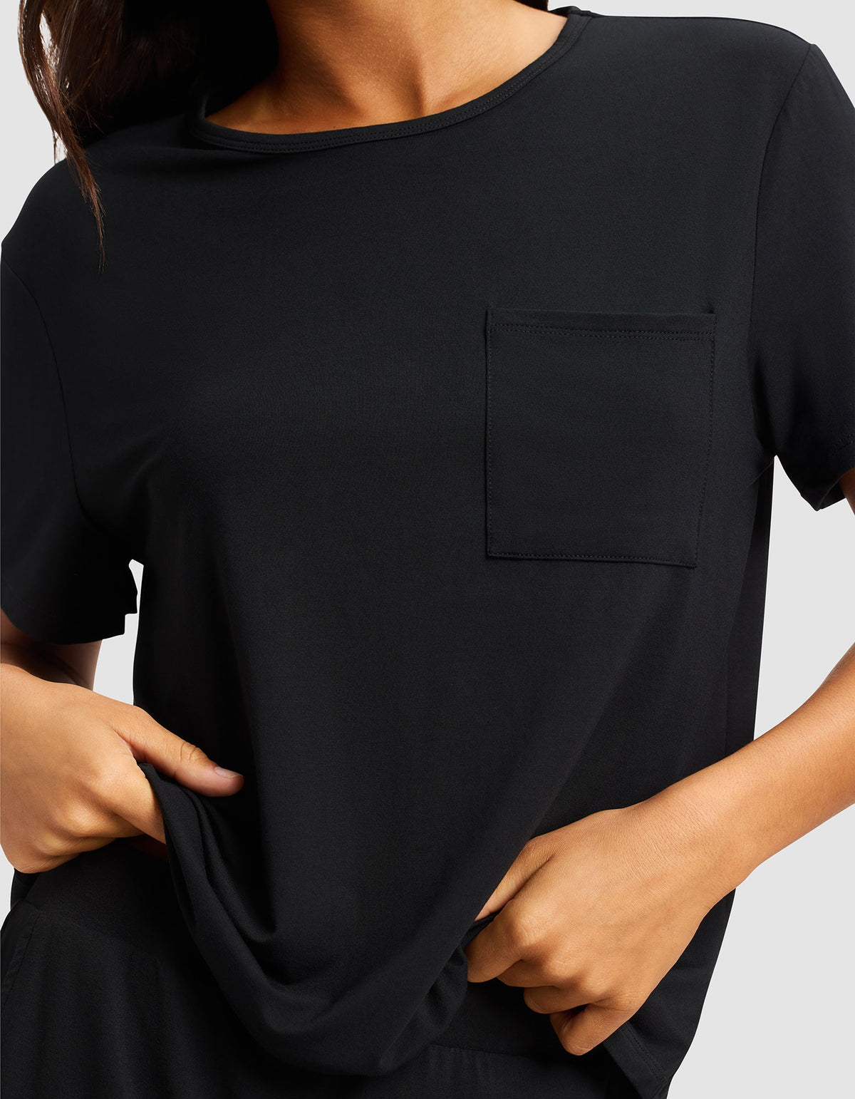 A woman is shown from shoulders to waist wearing the Cozy Earth Women's Bamboo Stretch-Knit Lounge Tee, a plain black short-sleeve shirt with a chest pocket, gently pulling the hem with both hands.|Color:Black