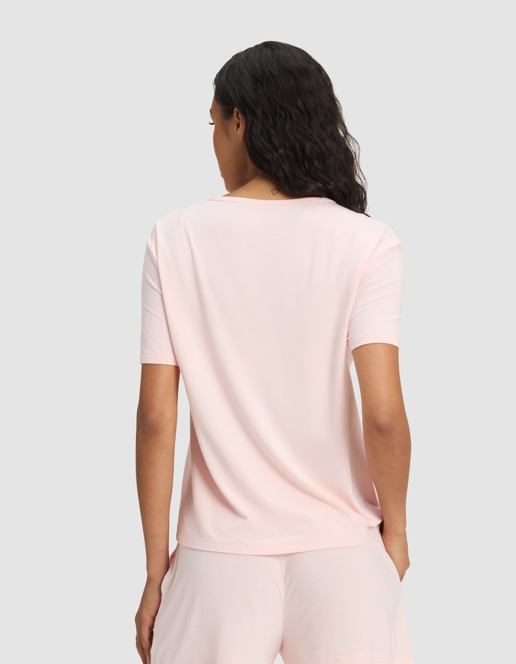 A woman with wavy dark hair is seen from the back wearing the Cozy Earth Women's Bamboo Stretch-Knit Lounge Tee & Pant Set in light pink, against a plain, light gray background.|Color:Blossom