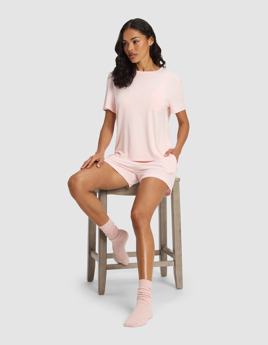 A woman with dark wavy hair sits on a wooden stool, wearing Cozy Earth’s Women's Bamboo Stretch-Knit Lounge Tee in light pink, matching shorts, and pink socks. She looks to the side with one leg on the stool and the other foot on the floor.|Color:Blossom