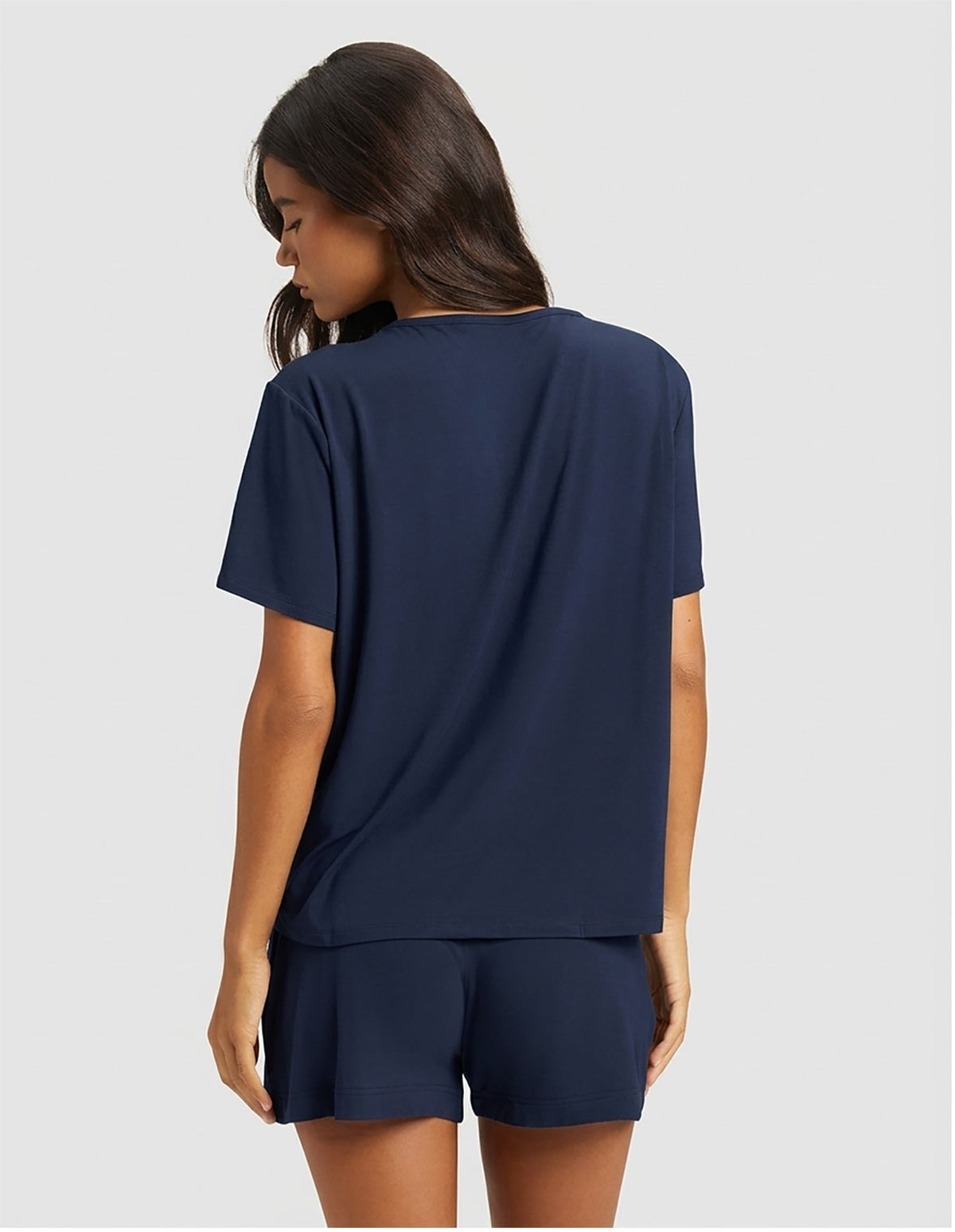 A woman with long dark hair stands facing away in a Cozy Earth Women's Bamboo Stretch-Knit Lounge Tee & Short Set, featuring a loose-fitting navy blue short-sleeve top and matching shorts against a plain light gray background.|Color:Navy