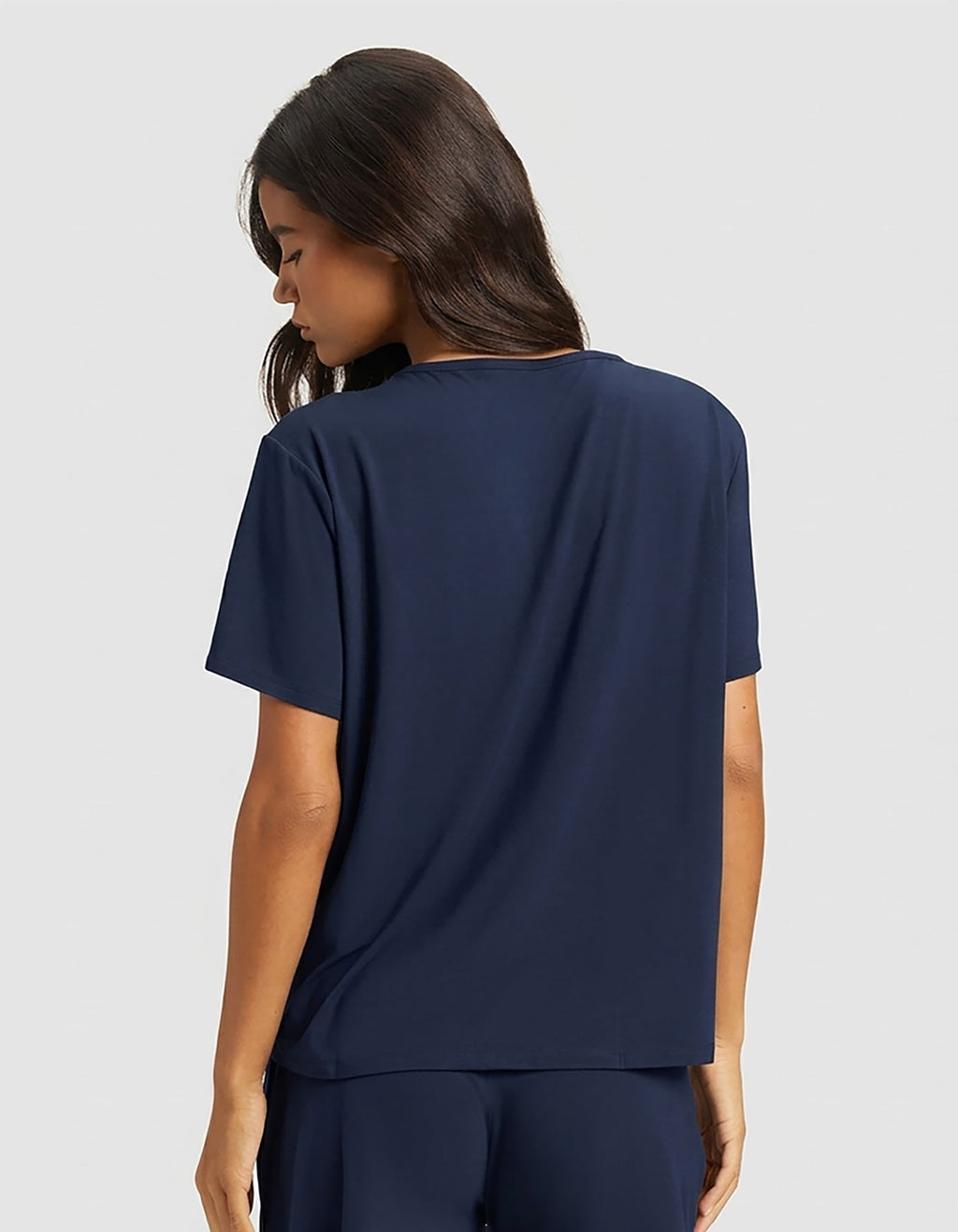 A woman with long dark hair models the Cozy Earth Women's Bamboo Stretch-Knit Lounge Tee & Pant Set in navy, standing with her back to the camera against a plain light gray background.|Color:Navy
