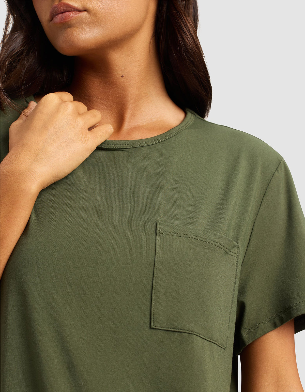 A woman models the Cozy Earth Women's Bamboo Stretch-Knit Lounge Tee in olive green, featuring short sleeves and a chest pocket, with one hand near her collarbone against a plain light gray background.|Color:Olive