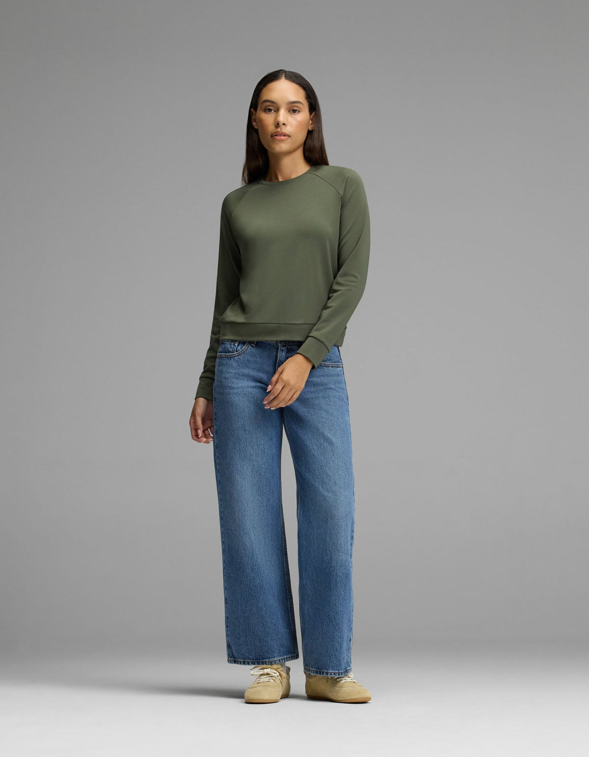 A woman stands against a gray background, wearing the Cozy Earth Women’s Brushed Bamboo Pullover Crew in olive green, paired with loose blue jeans and beige shoes. She has straight dark hair and a neutral expression, with her arms relaxed by her sides.|Color:Olive