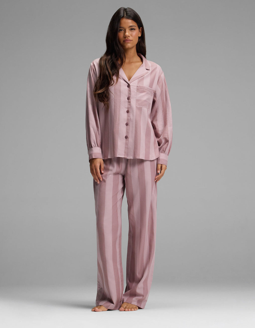 A woman stands barefoot, facing forward against a plain gray background, wearing the Cozy Earth Women’s Belmont Long Sleeve Pajama Set in Viola Stripe. The loose-fitting, button-up pajamas feature long sleeves and matching pants with a vertical stripe pattern.|Color:Viola Stripe
