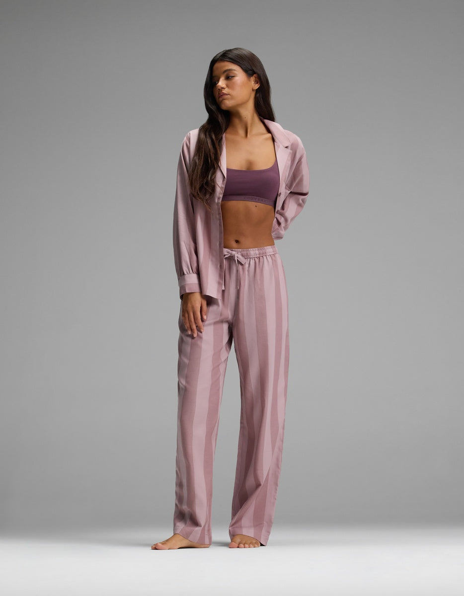 A woman stands barefoot against a plain gray background, wearing the Cozy Earth Women’s Belmont Pajama Pant in a pink striped design paired with a matching open shirt, revealing a purple sports bra underneath.|Color:Viola Stripe