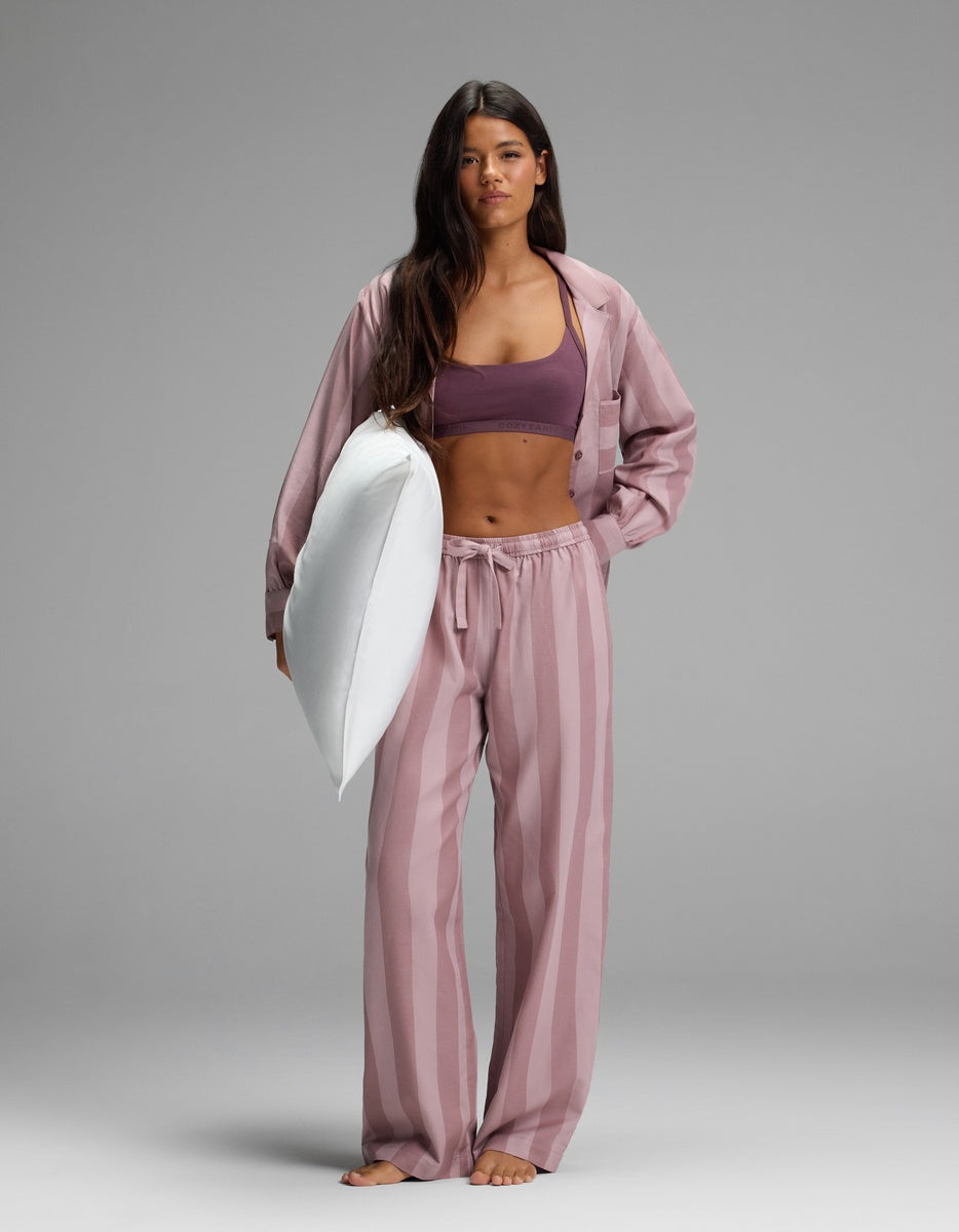 A woman wearing Cozy Earth’s Women’s Belmont Pajama Pant and a purple sports bra stands barefoot on a gray background, holding a white pillow under her arm and looking directly at the camera.|Color:Viola Stripe