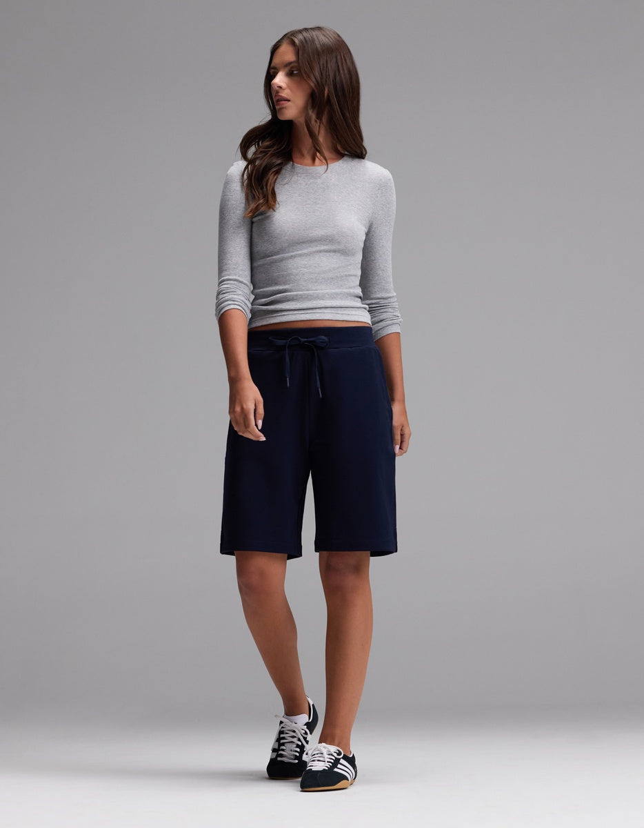 A woman stands against a plain gray background wearing a fitted gray long-sleeve top, black sneakers, and Cozy Earth’s Women’s Brushed Bamboo Bermuda Shorts in navy with a drawstring waist.|Color:Navy