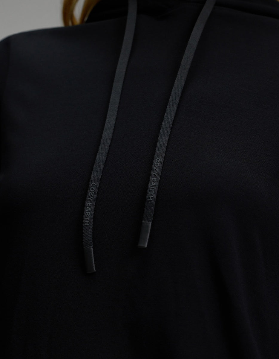 Close-up of the Women’s Brushed Bamboo Hoodie by Cozy Earth in black, highlighting the chest area and two black drawstrings with “COZY EARTH” embossed on the tips.|Color:Black