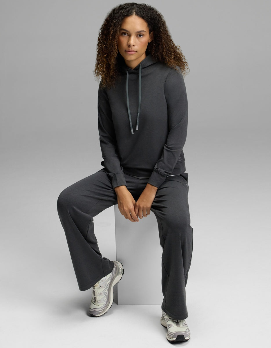 A woman with curly hair wears the Cozy Earth Women’s Brushed Bamboo Hoodie paired with matching pants, sitting on a white block and looking at the camera. She completes the look with white sneakers, set against a plain light gray background.|Color:Charcoal