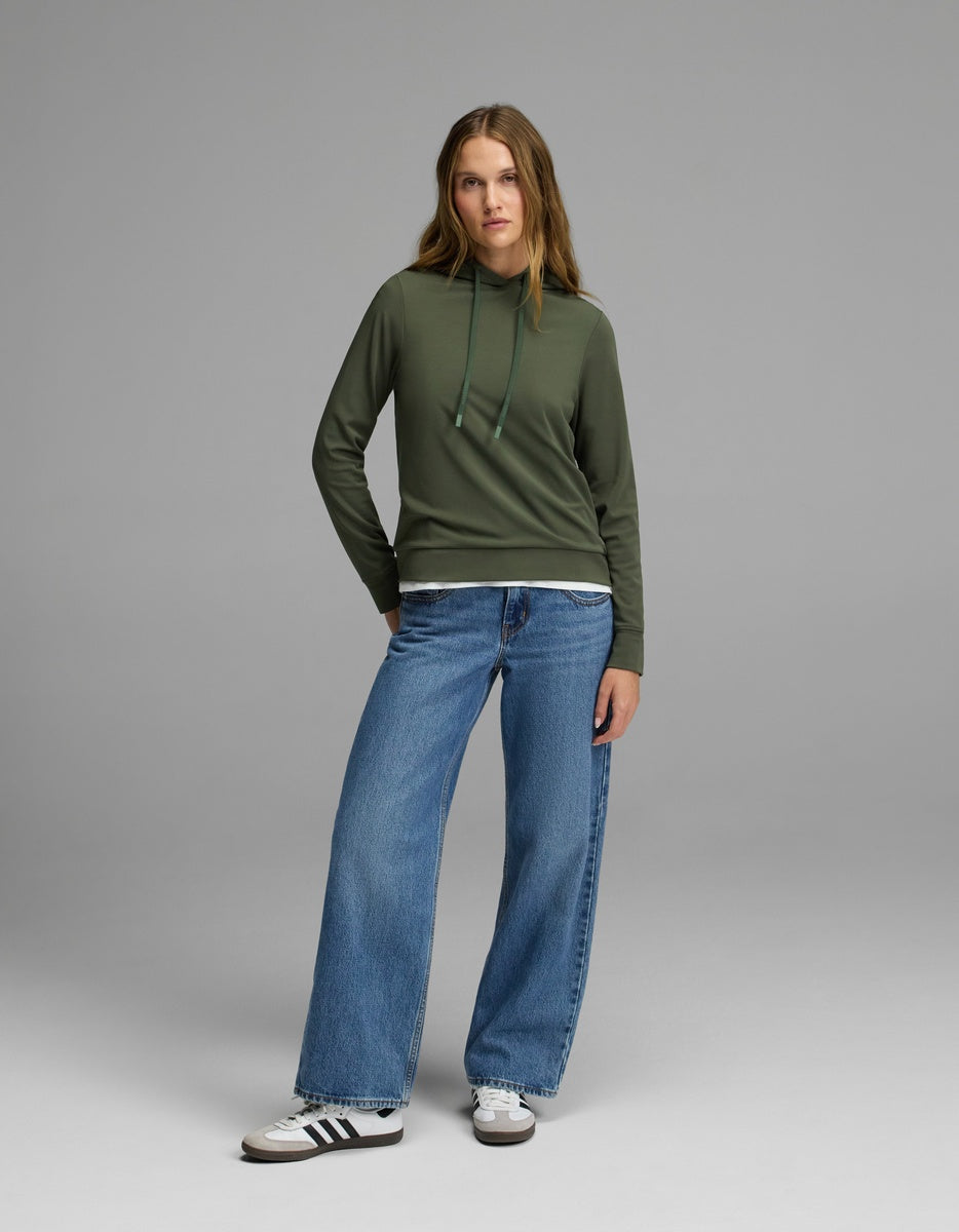 A woman stands facing forward against a plain gray background, wearing the Cozy Earth Women’s Brushed Bamboo Hoodie in green, paired with blue wide-leg jeans and white Adidas sneakers with black stripes.|Color:Olive