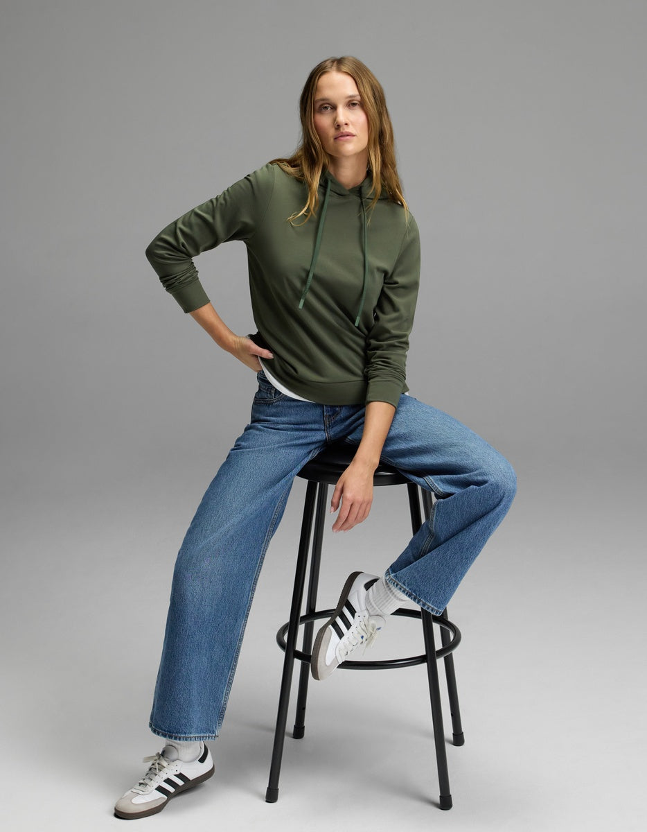 A person wearing the Cozy Earth Women’s Brushed Bamboo Hoodie, blue jeans, and white Adidas sneakers sits on a black stool against a plain gray background.|Color:Olive