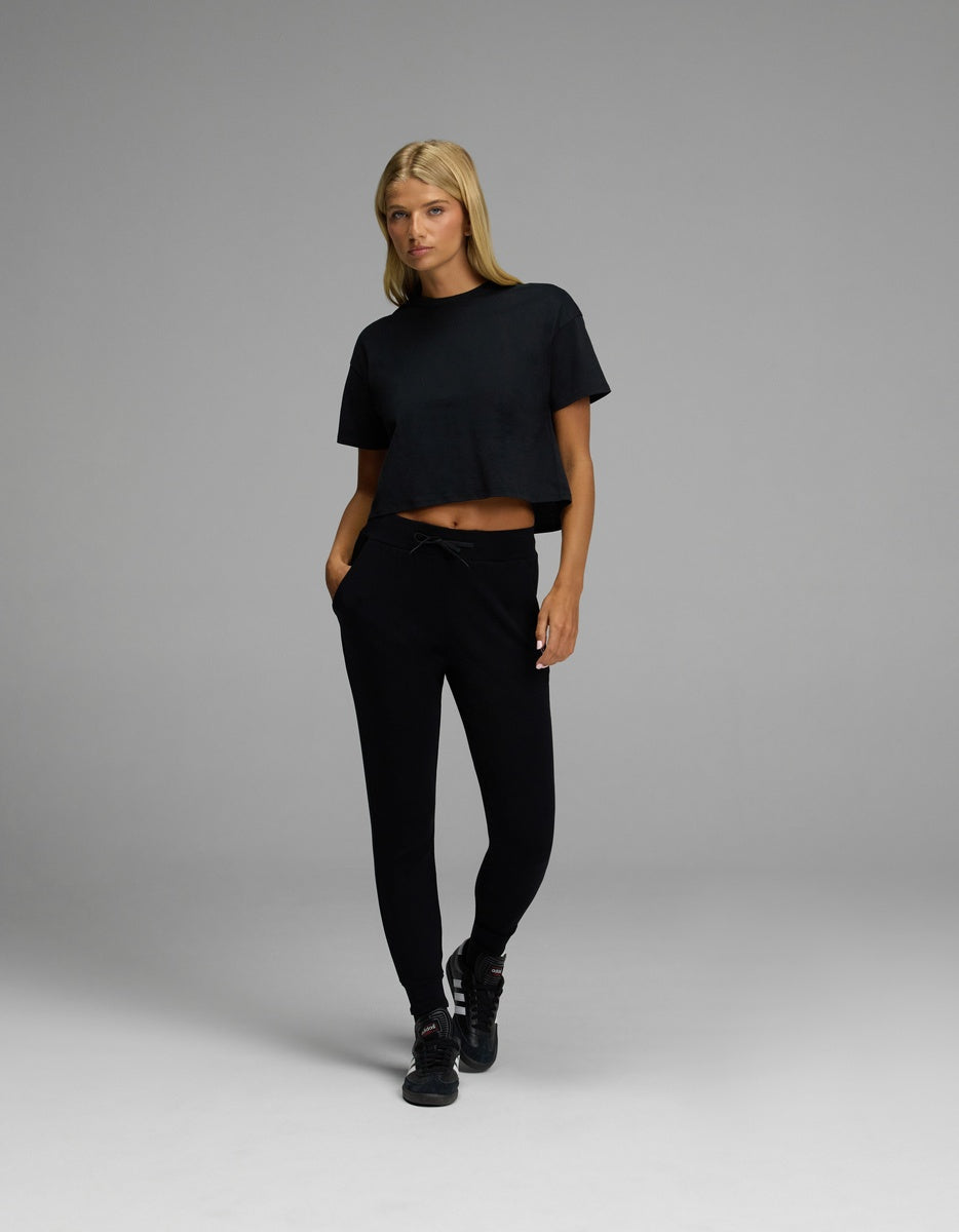 A person with long blonde hair stands with one foot forward and hands in pockets against a plain gray background, wearing black sneakers, a black cropped t-shirt, and Cozy Earth’s Women’s Brushed Bamboo Jogger.|Color:Black