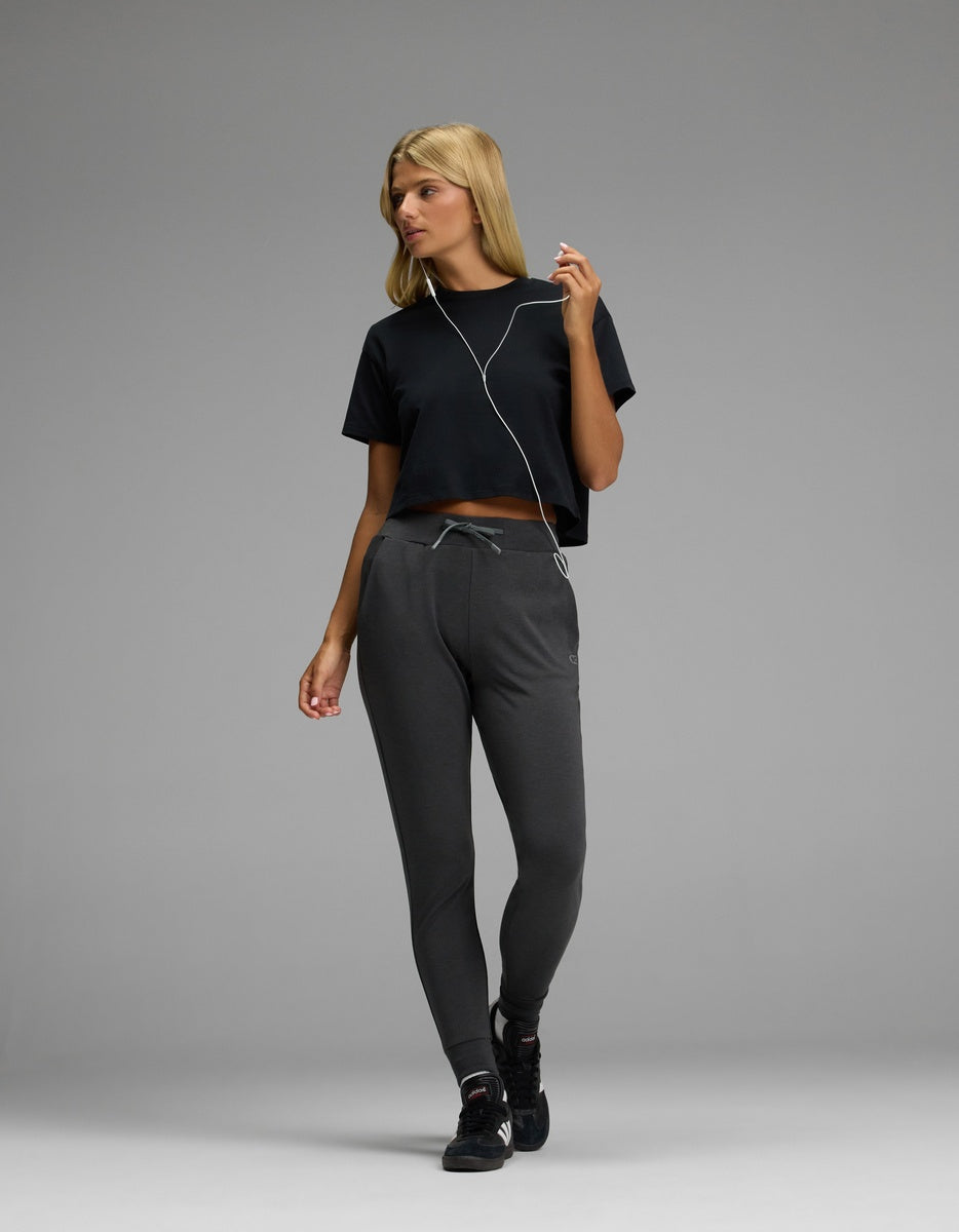 A woman with blonde hair is standing against a plain gray background, looking to the side while listening to earphones. She is wearing a black cropped t-shirt, black sneakers, and the Cozy Earth Women’s Brushed Bamboo Jogger in dark gray.|Color:Charcoal