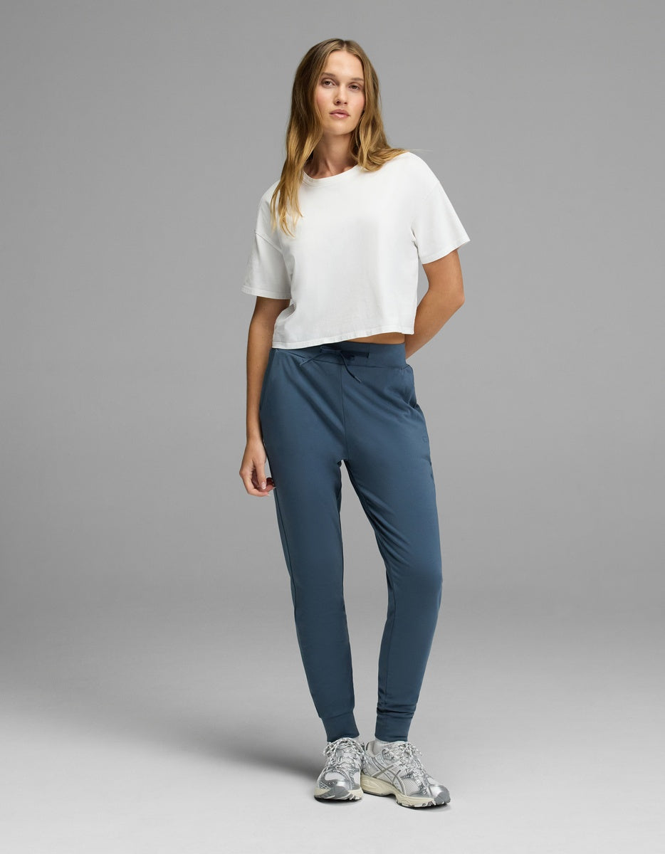 A woman stands facing forward against a plain gray background, wearing Cozy Earth’s Women’s Brushed Bamboo Jogger in blue, paired with a white cropped t-shirt and gray sneakers.|Color:Dawn