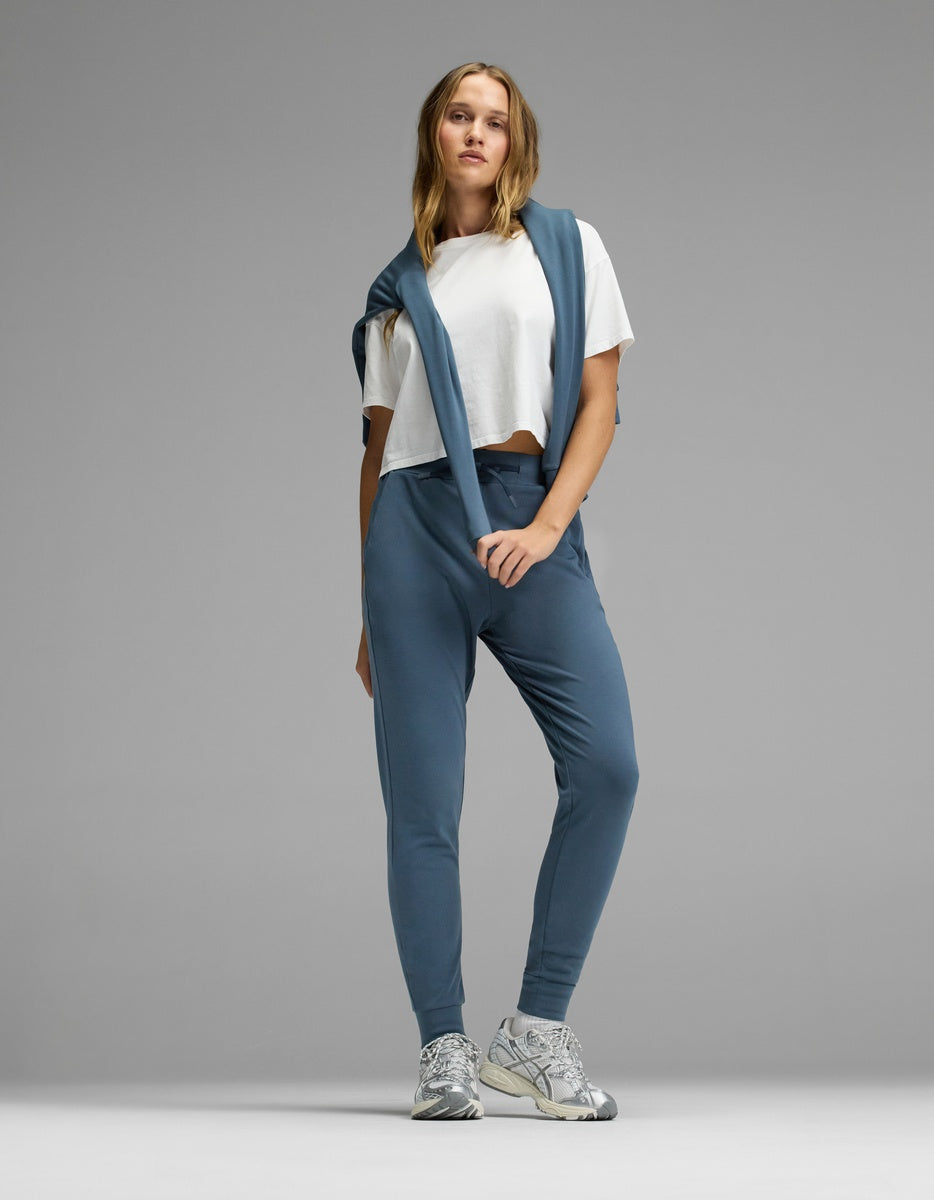 A person stands against a plain gray background in casual attire, featuring the Cozy Earth Women’s Brushed Bamboo Jogger in blue, a white cropped T-shirt, a matching blue sweatshirt draped over their shoulders, and silver sneakers.|Color:Dawn