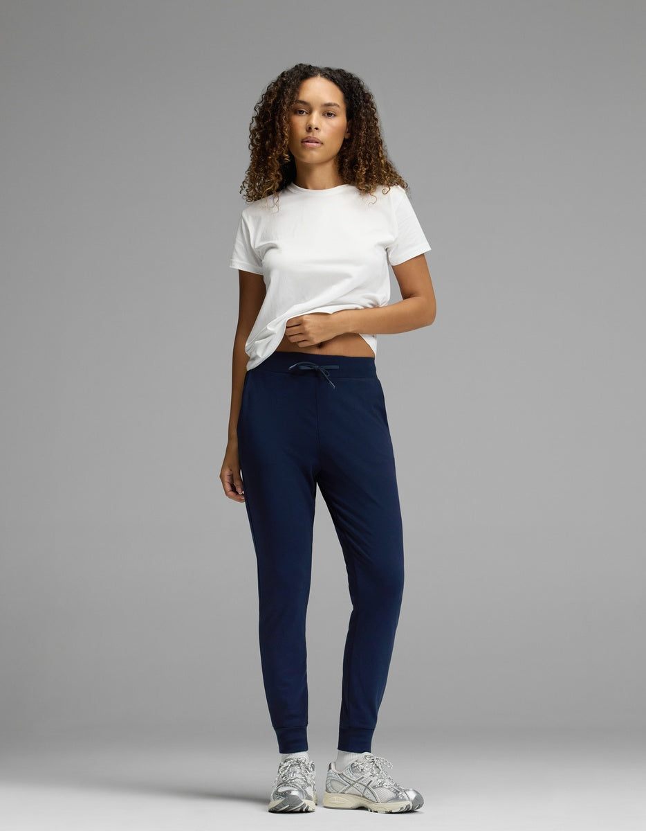A woman stands against a plain gray background, wearing Cozy Earth’s Women’s Brushed Bamboo Jogger in dark blue, paired with a white t-shirt and gray sneakers. Her curly hair is down and she has one hand in her pocket.|Color:Navy