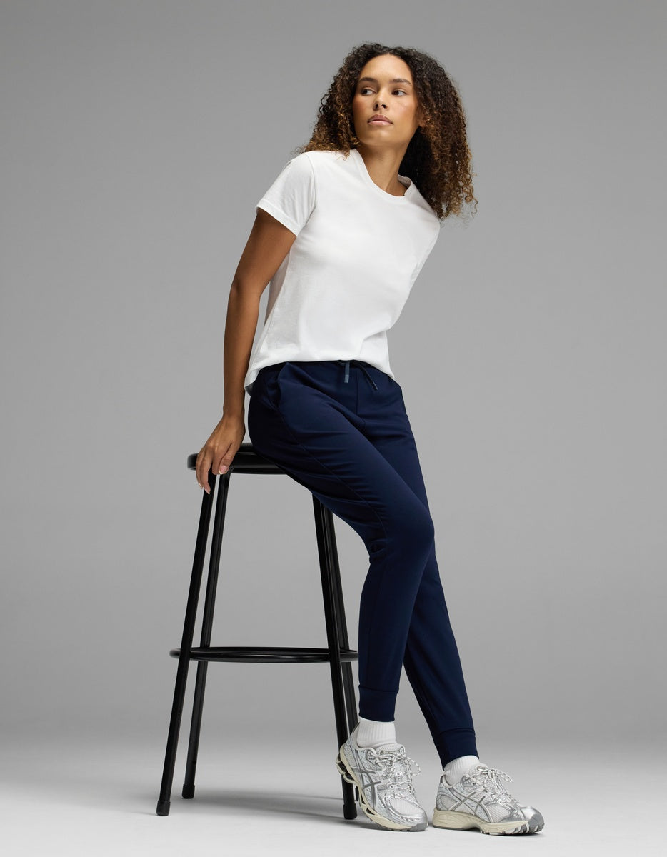 A woman wearing Cozy Earth’s Women’s Brushed Bamboo Jogger in navy, paired with a white t-shirt and gray sneakers, sits sideways on a tall black stool against a plain gray background, looking to her left.|Color:Navy