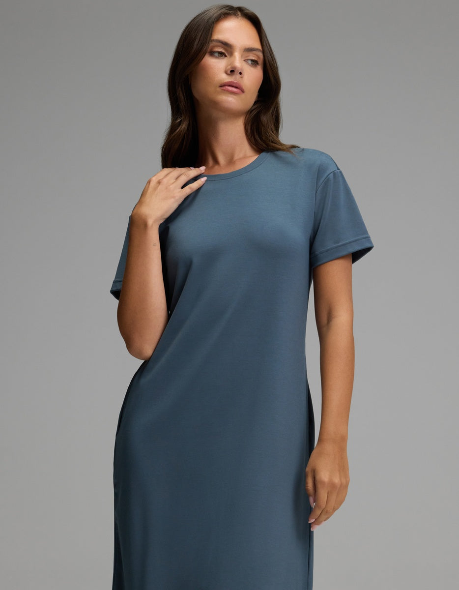 A woman with long brown hair models the Cozy Earth Women’s Brushed Bamboo Midi Dress in slate blue, featuring a loose fit and short sleeves, as she poses against a plain gray background, gazing slightly to the side with one hand gently resting near her collarbone.|Color:Dawn