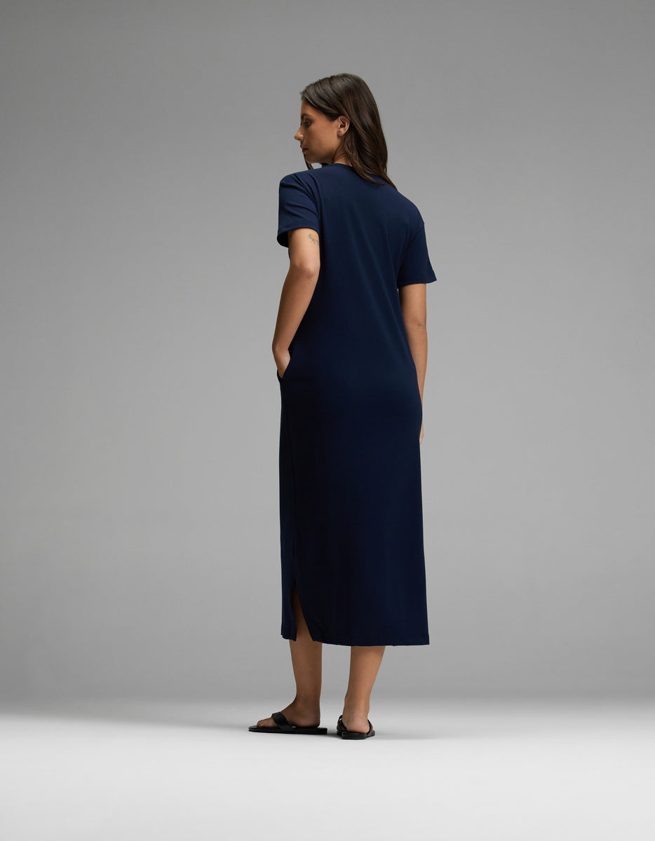 A woman stands against a plain background, facing away, wearing the Cozy Earth Women’s Brushed Bamboo Midi Dress in navy blue with short sleeves, side slits, and black sandals.|Color:Navy