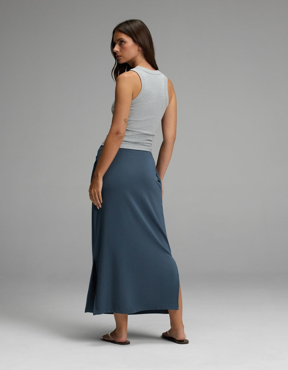 A woman stands with her back to the camera, wearing a sleeveless gray top and the Cozy Earth Women’s Brushed Bamboo Midi Skirt in blue, featuring a side slit. She pairs the outfit with sandals and glances over her shoulder against a plain gray background.|Color:Dawn